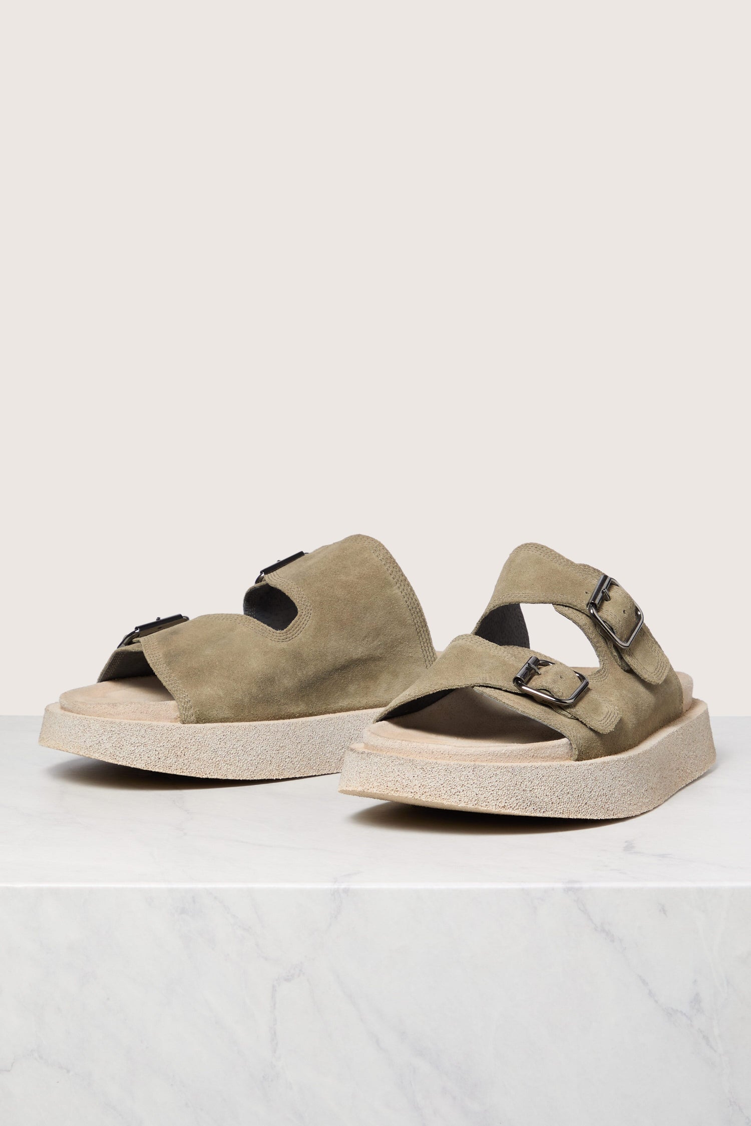 A pair of Italian construction Suede Double Buckle Slider sandals on a white surface against a marble backdrop.