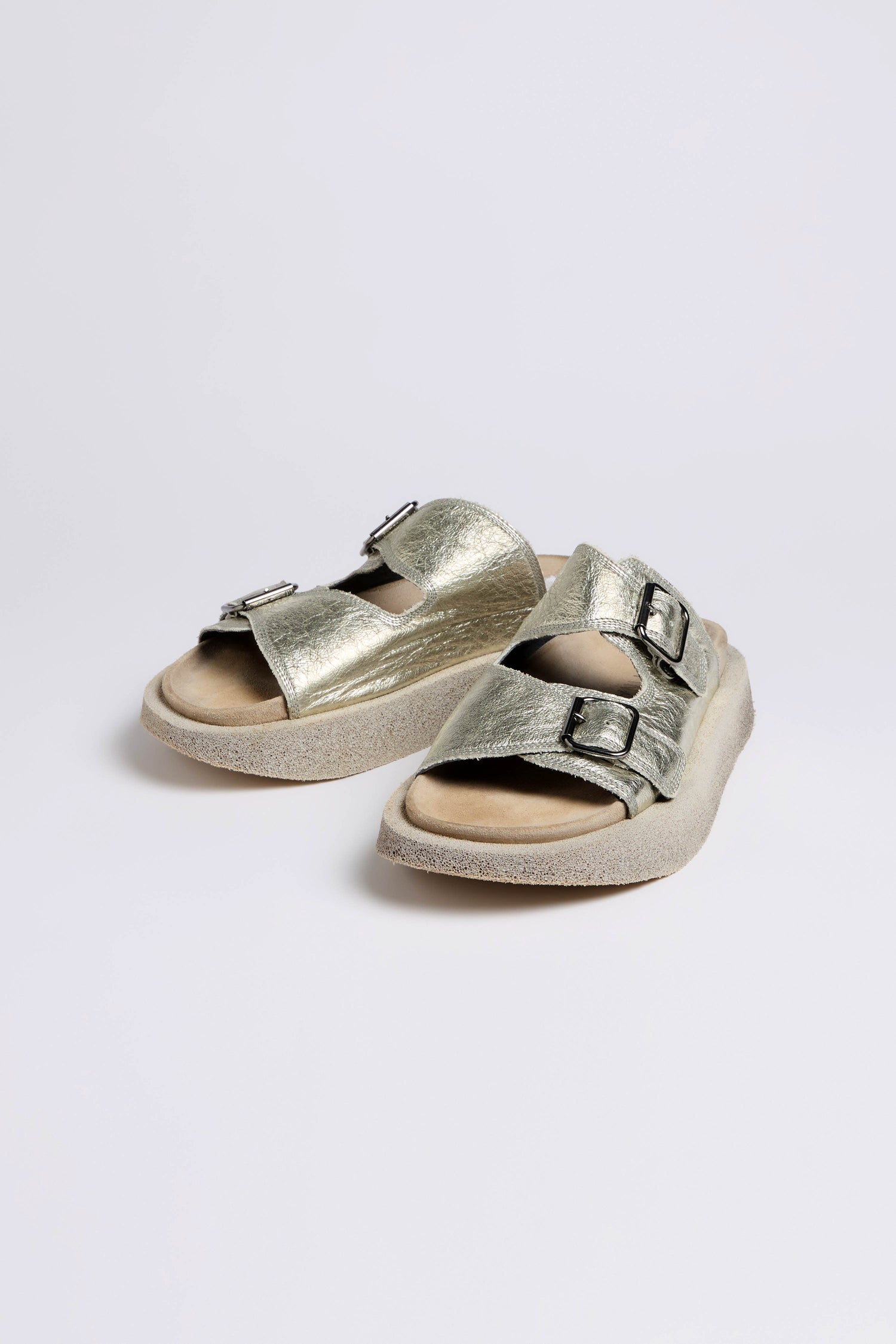 A pair of Metallic Silver Double Buckle Leather Sliders with adjustable straps and cushioned soles rest on a light gray surface against a white background.