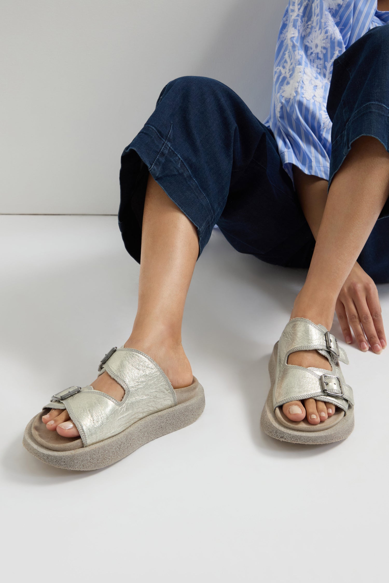 A person sits cross-legged, highlighting handcrafted Italian footwear—the Metallic Silver Double Buckle Leather Sliders. Dark pants and a blue striped shirt perfectly complement the chic sliders.