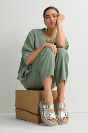 A woman elegantly sits on stacked wooden blocks in a textured green outfit and Metallic Silver Double Buckle Leather Sliders, her hand thoughtfully resting on her face against a plain backdrop.