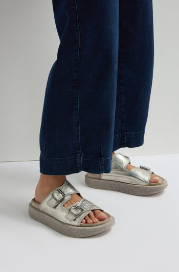 A person wearing blue jeans and Metallic Silver Double Buckle Leather Sliders stands on a white background.