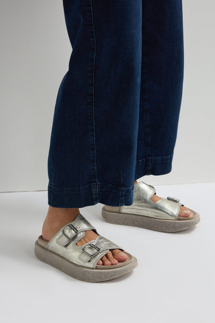 A person wearing blue jeans and Metallic Silver Double Buckle Leather Sliders stands on a white background.