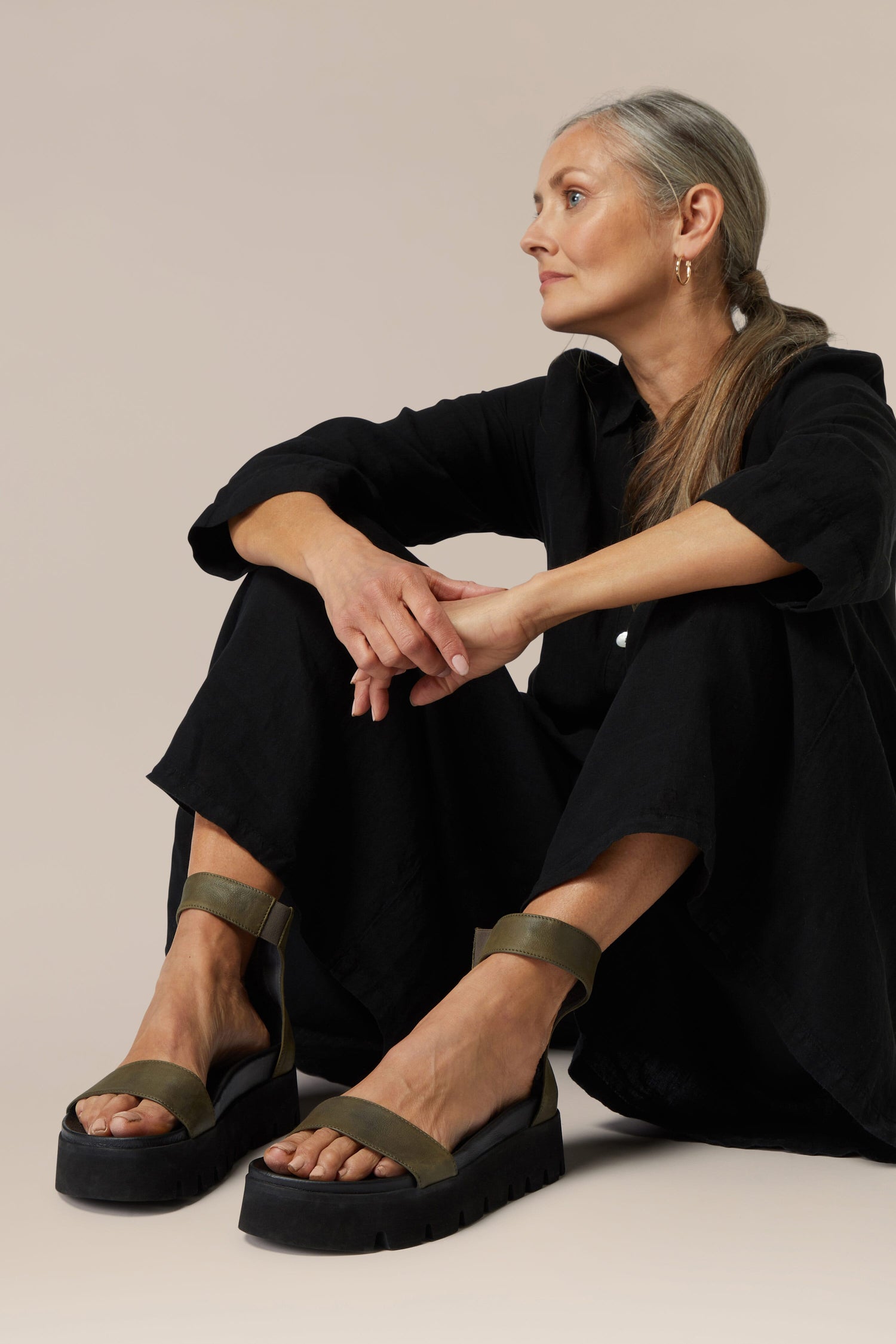 A woman in a black outfit and green Leather Platform Zip Sandal, featuring handmade Italian construction, sitting against a neutral background.