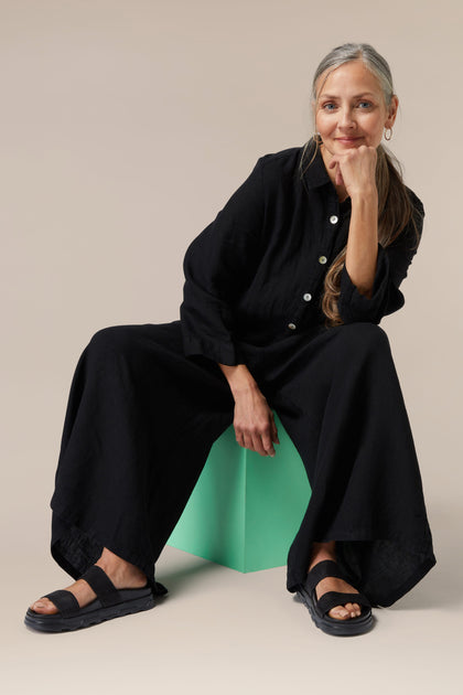 Sentence with Product Name: A woman sitting relaxed on a stool, wearing a black Fabletics Stretch Leather Slider shirt and wide-leg pants, with a contemplative expression.