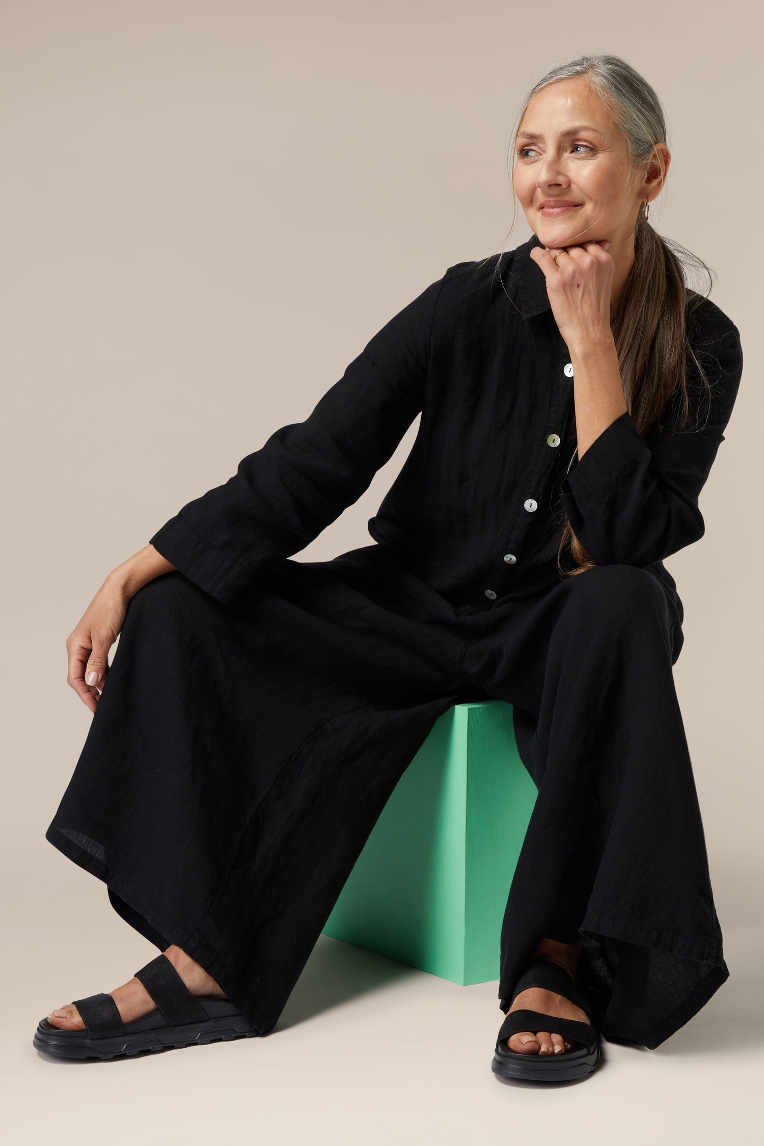 A woman in a black Stretch Leather Slider outfit sitting on a green stool with one hand resting on her chin, looking thoughtfully to the side.