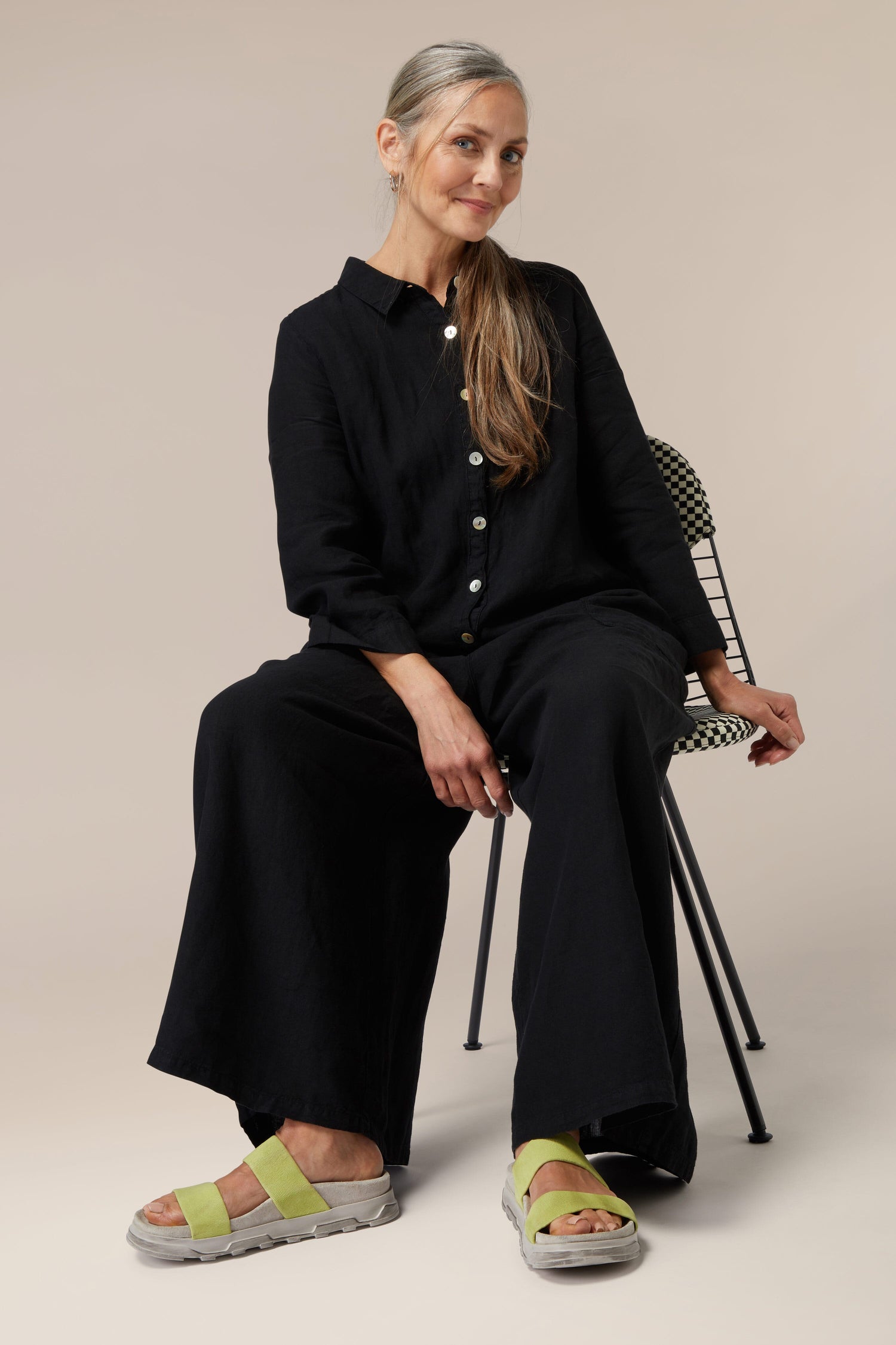 A woman in a black outfit and Stretch Leather Slider sitting on a chair against a neutral background.