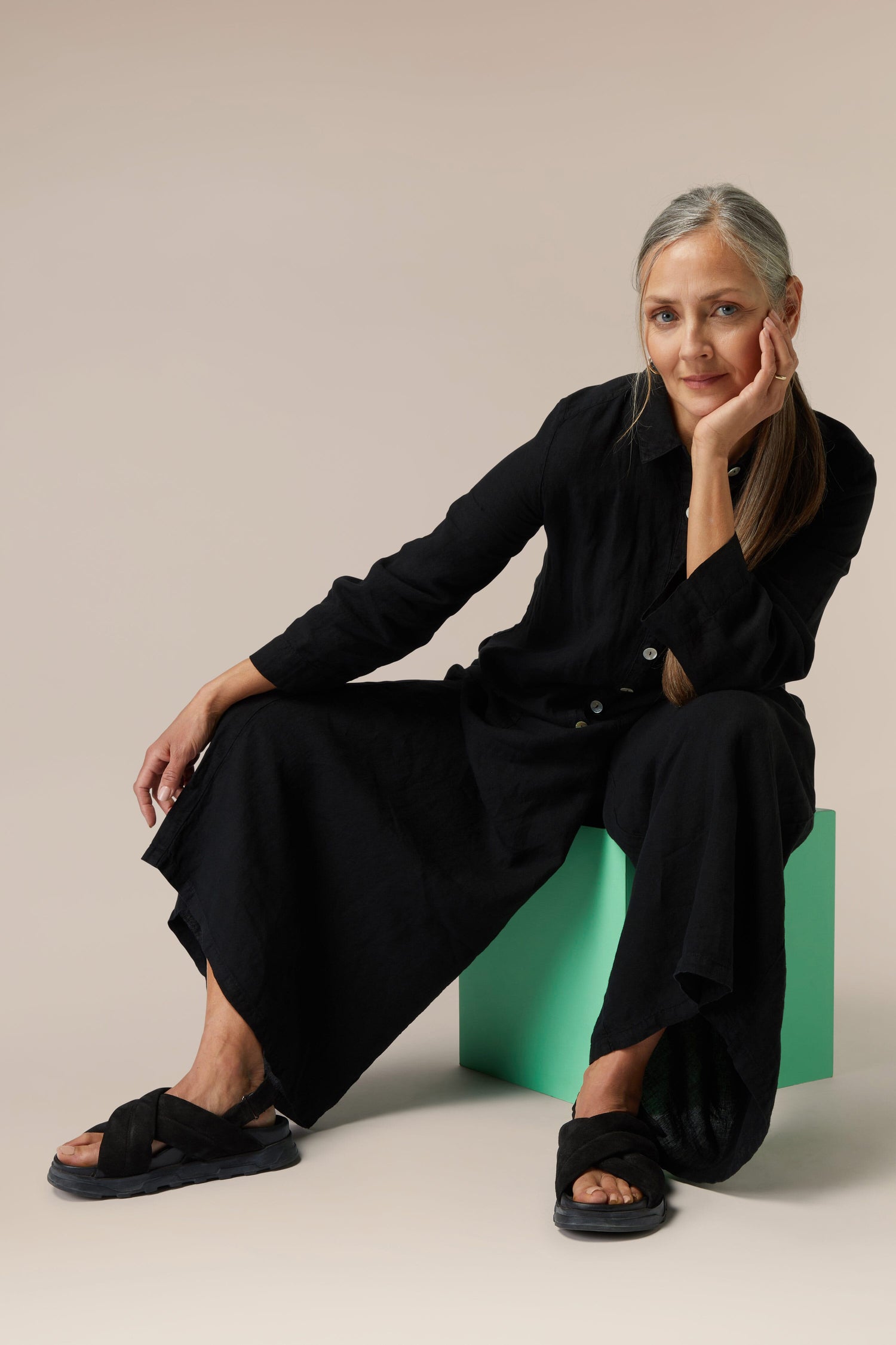 Woman sitting on a green stool, wearing a black outfit and Suede Crossover Sandals.