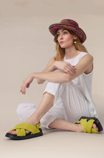 Woman sitting on the floor wearing a white outfit, colorful hat, and yellow Suede Crossover Sandal.