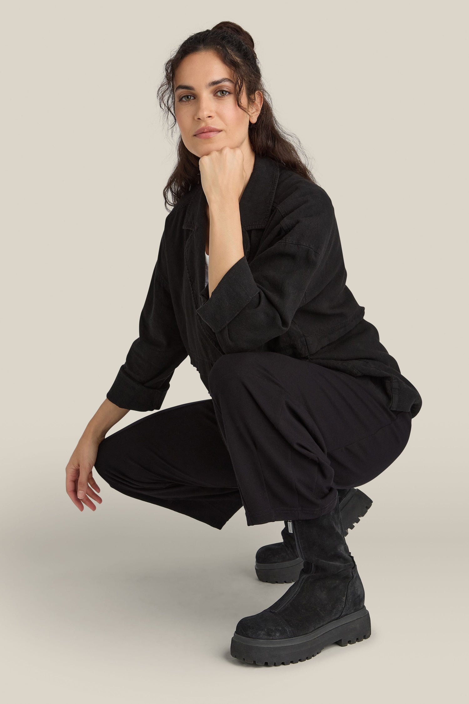 A person with long dark hair, wearing a black jacket, black pants, and Zip Front Suede Platform Boots, is squatting against a plain neutral background.