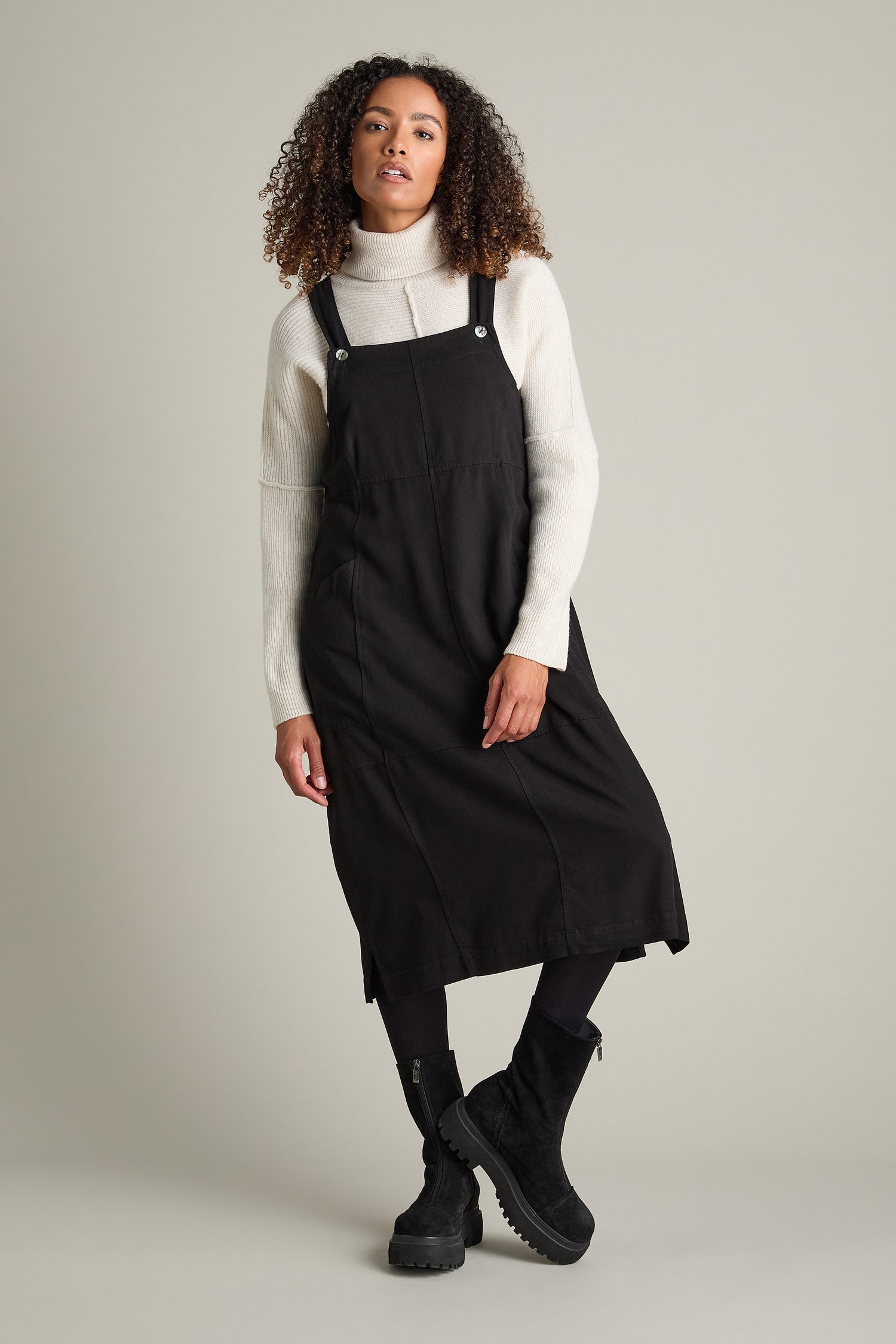 A person stands against a gray background, wearing a black pinafore dress over a cream turtleneck sweater, black tights, and Zip Front Suede Platform Boots. The person has curly hair and a neutral expression.