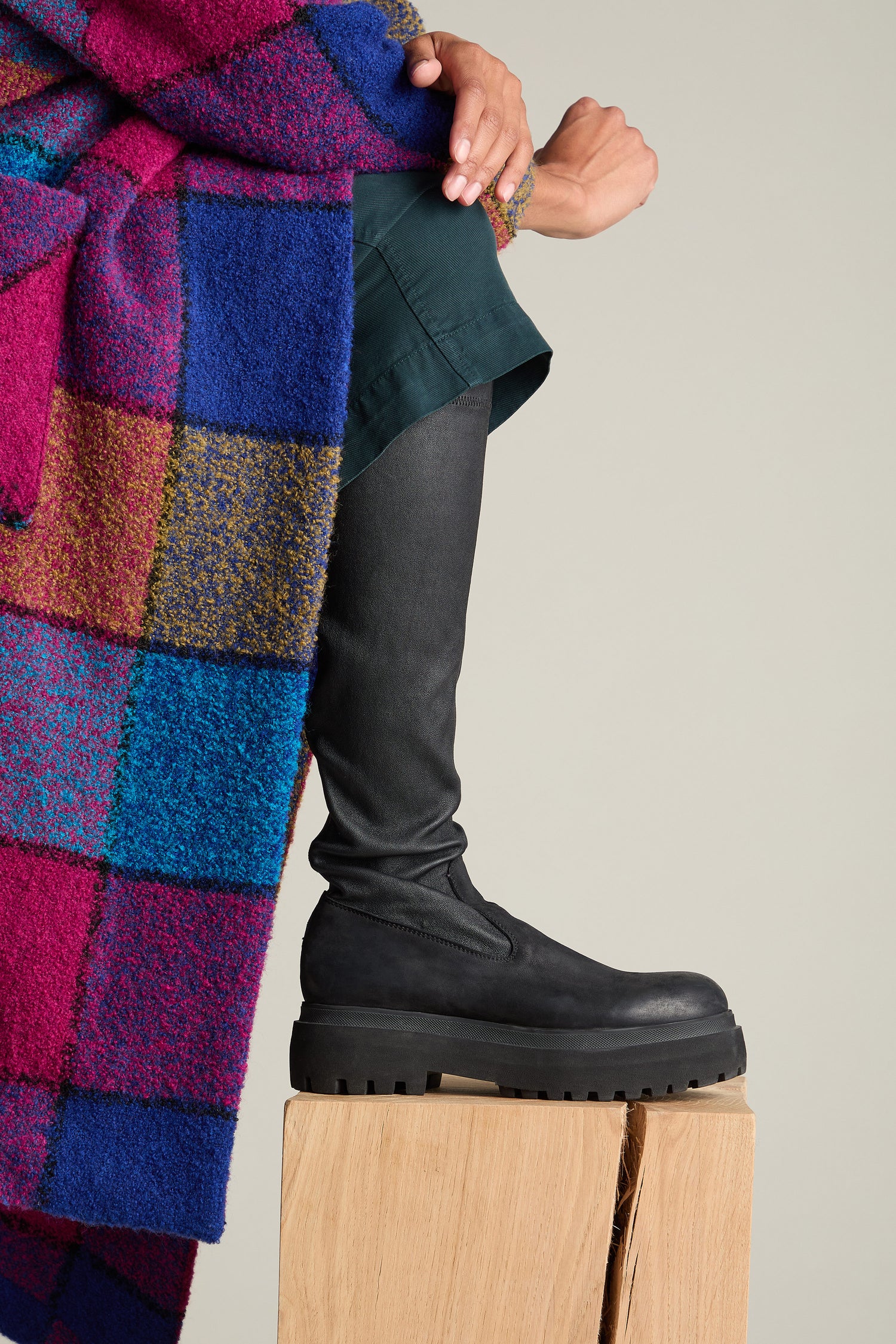 A person wearing a colorful checkered blanket and black Long Stretch Knee High Leather Boots sits with one leg crossed over the other on a wooden block.