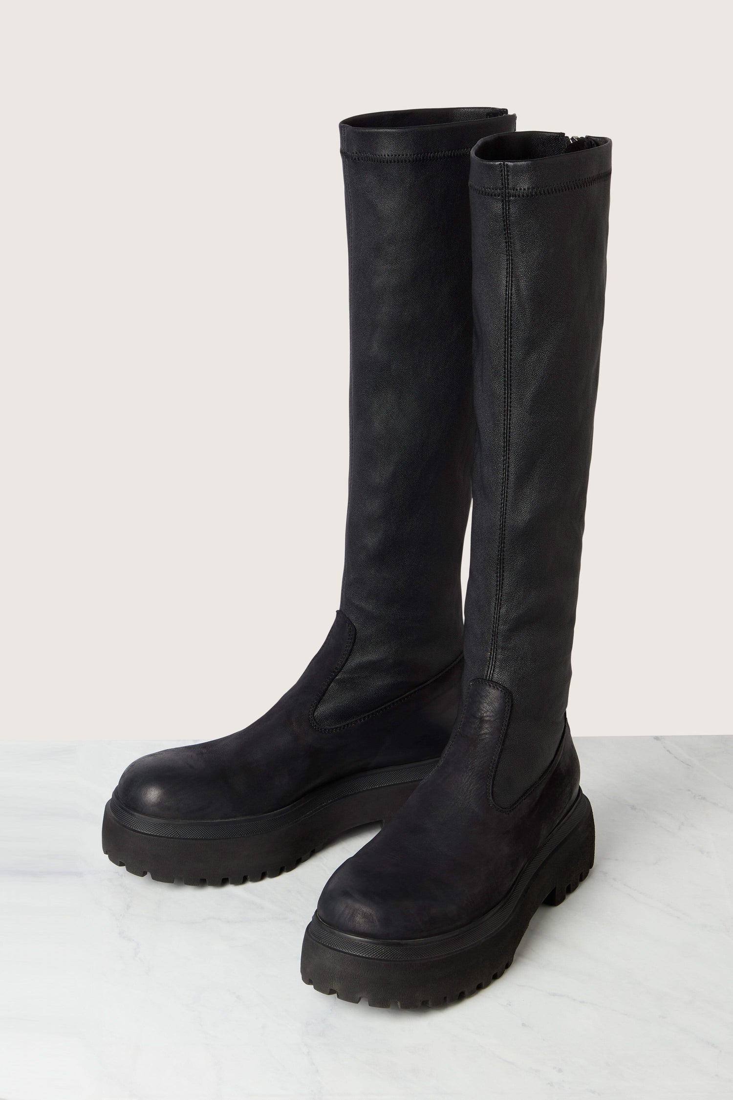 A pair of Long Stretch Knee High Leather Boots, featuring a tall design, black leather construction, rugged soles, and back zippers, are placed on a marble surface against a light background.