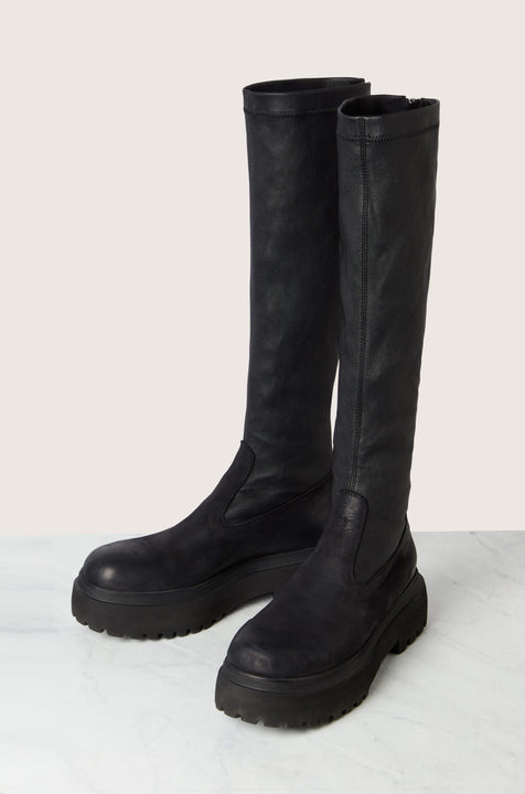 A pair of Long Stretch Knee High Leather Boots, featuring a tall design, black leather construction, rugged soles, and back zippers, are placed on a marble surface against a light background.