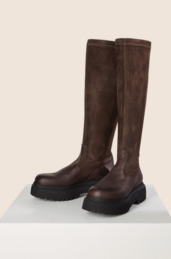 The Long Stretch Knee High Leather Boots, crafted from dark brown calfskin leather and featuring thick black rubber soles reminiscent of Lofina boots, are elegantly displayed on a white surface.