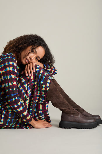 A person in a colorful geometric-patterned dress sits leaning on their knees, wearing tall Long Stretch Knee High Leather Boots against a plain background.