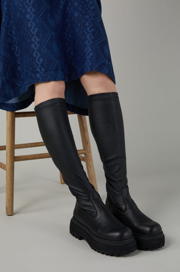 Person sitting on a wooden chair wearing a blue patterned skirt and striking Black Long Stretch Knee High Leather Boots with thick soles.