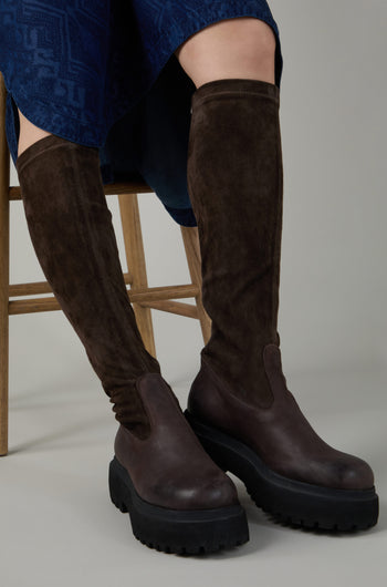 A person sits on a wooden chair, legs slightly apart, wearing Chocolate Long Stretch High Knee Suede Boots with a thick platform sole and a blue skirt visible.