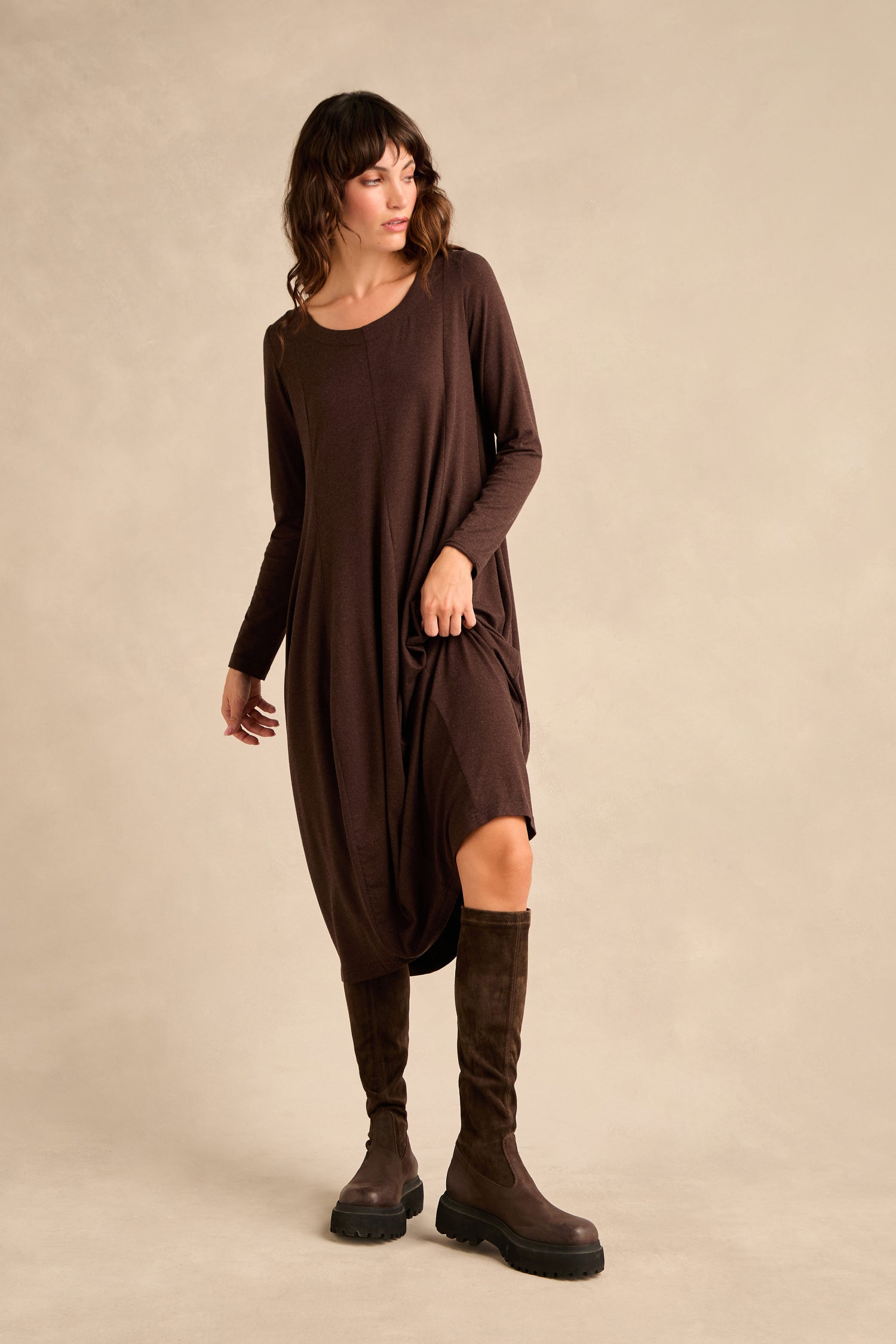 A woman stands against a neutral background, wearing knee high stretch suede boots and a long-sleeve brown dress with side pockets.