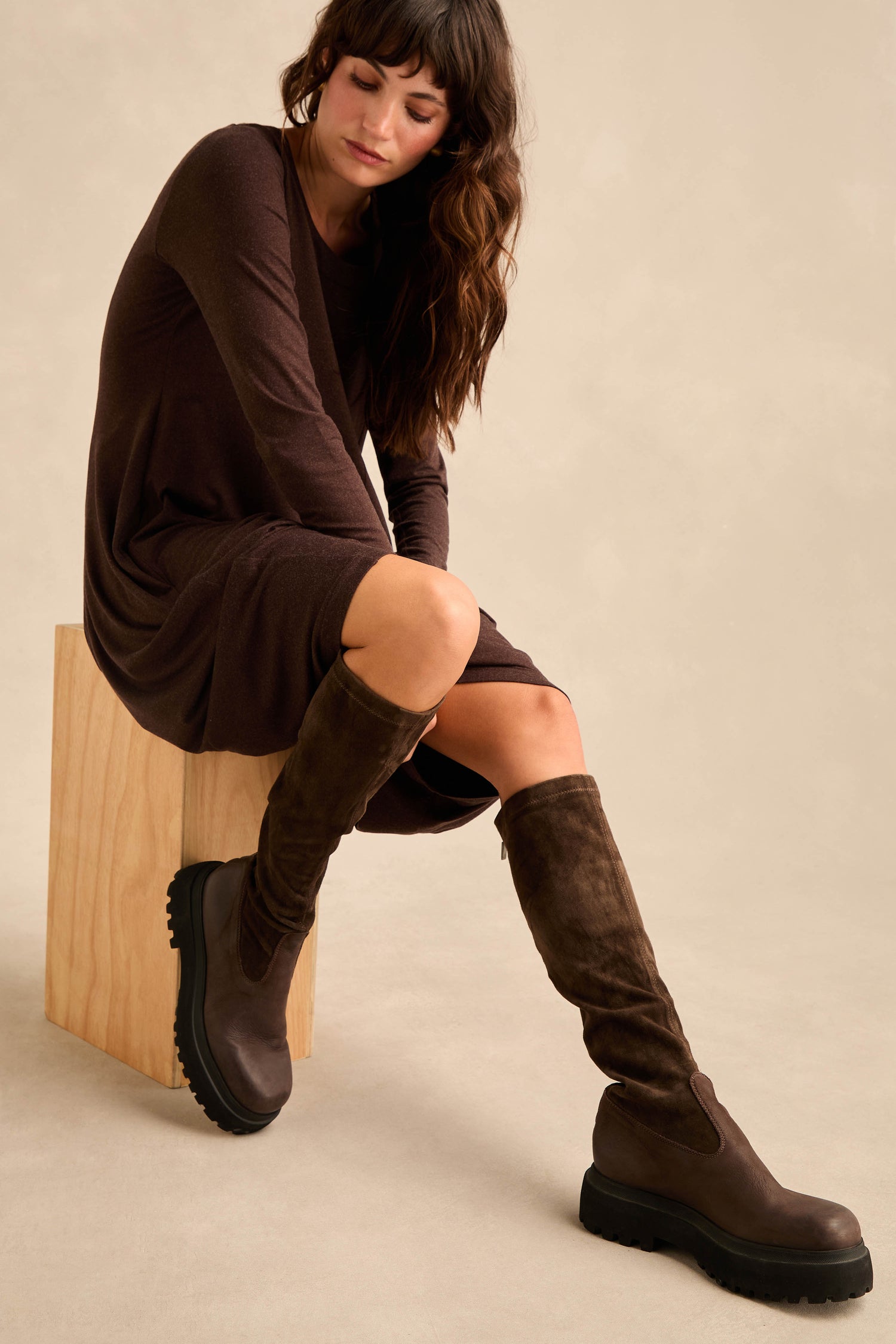 A woman wearing Knee High Stretch Suede Boots and a brown long-sleeve dress sits on a simple wooden block against a neutral background.