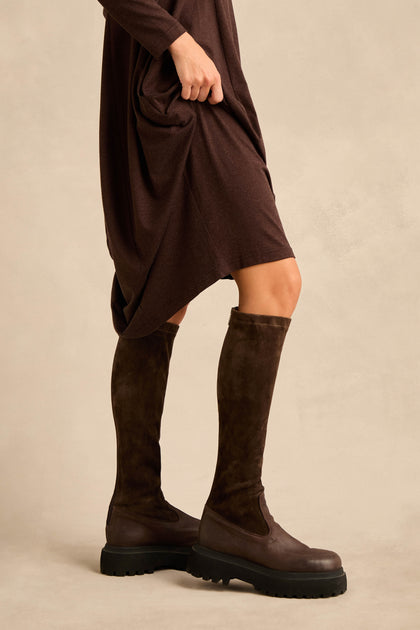 A person models the Knee High Stretch Suede Boots, featuring chunky black soles, paired with a brown dress against a neutral background.