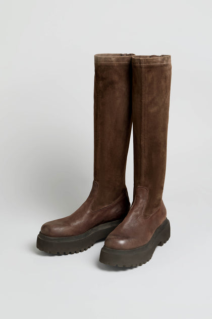 A pair of Knee High Stretch Suede Boots in a rich brown shade, featuring thick black rubber soles and rounded toes, positioned side by side on a plain light background.