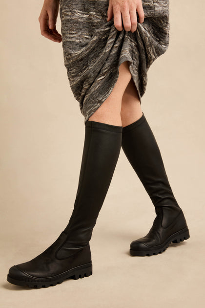 A person wearing a textured gray skirt and black Long Nappa Leather Stretch Boots stands on a beige background, with one hand lifting the skirt slightly.