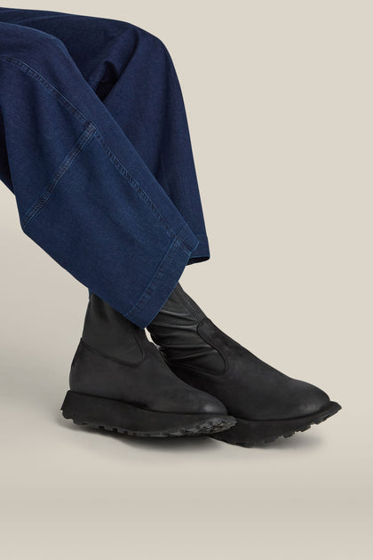 A person wearing dark blue wide-legged pants and black Leather Stretch Boots is seated against a beige background.