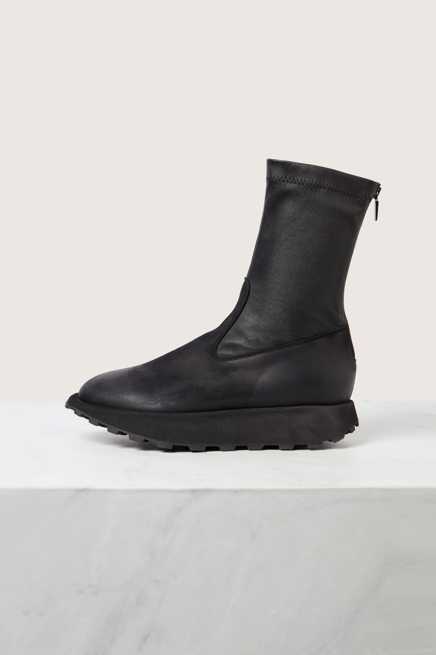 A Leather Stretch Boot by Lofina, featuring a black high-top design with a rugged sole and back zipper, crafted from supple calfskin, displayed on a white marble surface.