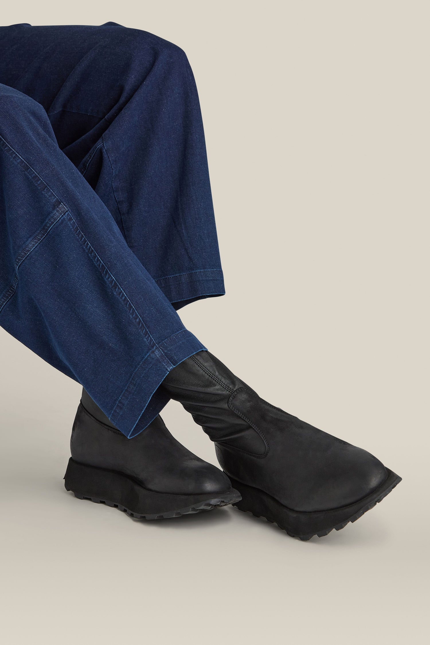 Close-up of a person wearing dark blue pants and the Leather Stretch Boot in sleek black leather against a beige background.