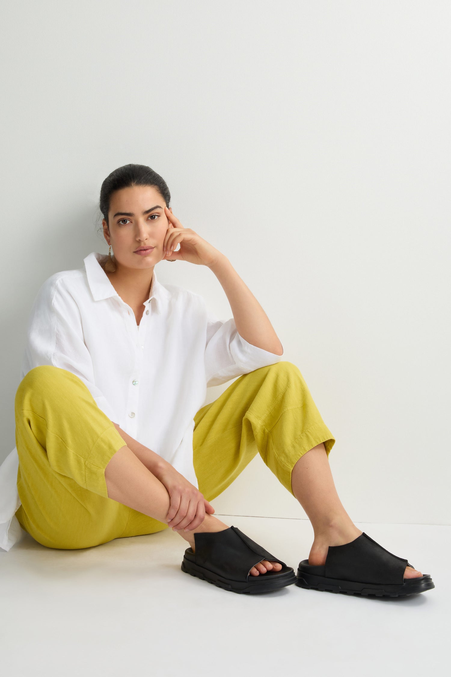 A person in a white shirt, yellow pants, and Seam Leather Sliders with a cushioned sole sits against a white wall on a wooden floor, embodying modern minimalism.