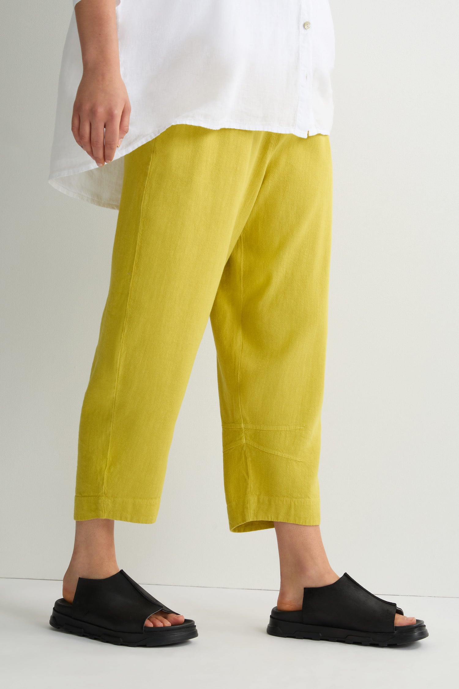 Against a plain backdrop, an individual in a white shirt and yellow pants wears Seam Leather Sliders with cushioned soles, epitomizing modern minimalism.