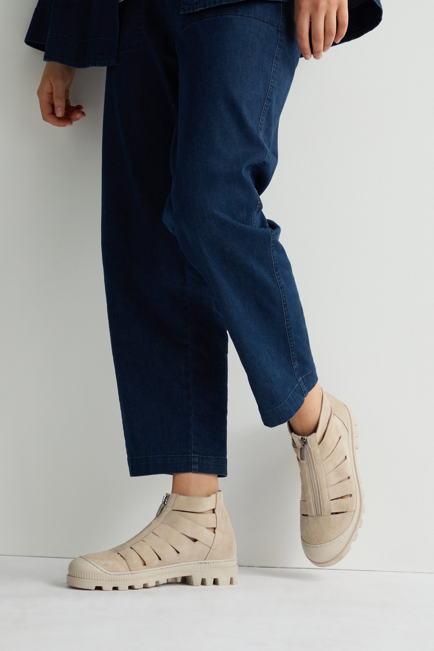 Against a white background, an individual in blue pants showcases edgy Cut Out Suede Zip Boots featuring rugged rubber soles.