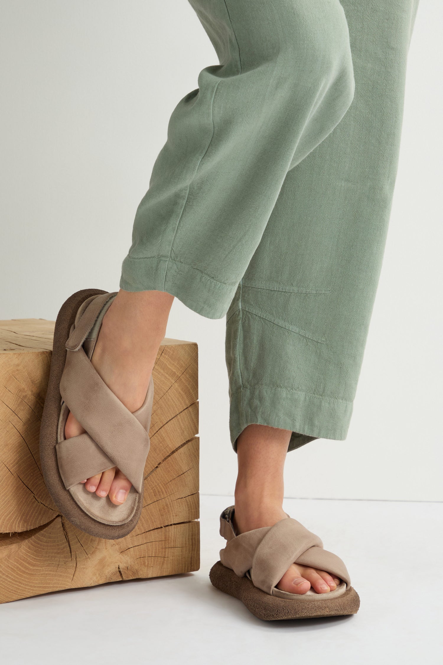 A person in green pants and beige Crossover Leather Sandals, renowned for their Scandinavian craftsmanship, steps on a wooden block.