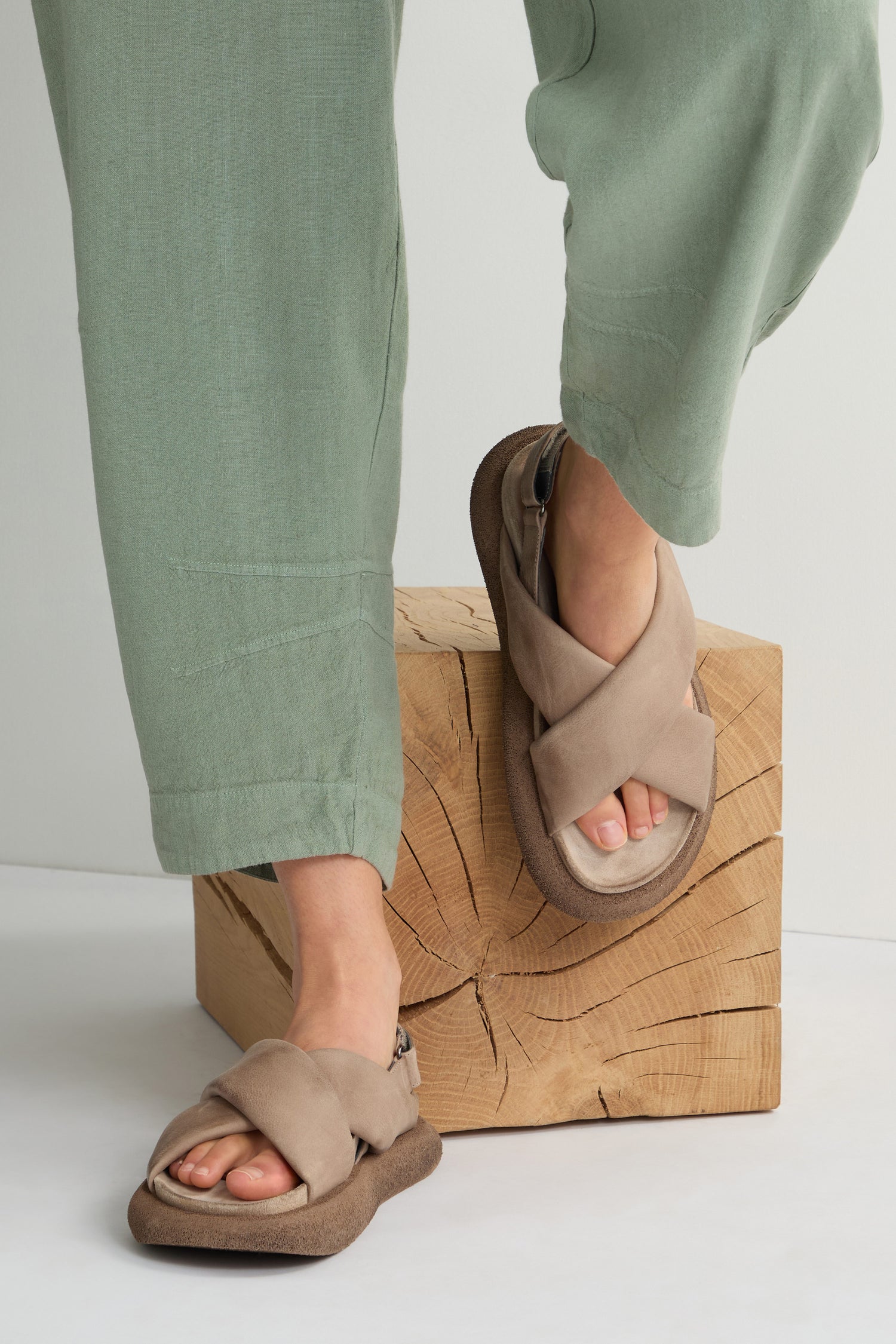 A person wearing green pants and Crossover Leather Sandals, renowned for their Scandinavian craftsmanship, stands on a wooden block.