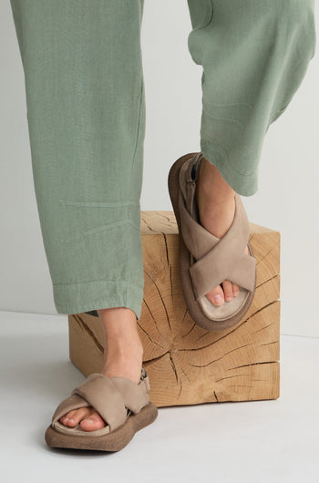 A person wearing green pants and Crossover Leather Sandals, renowned for their Scandinavian craftsmanship, stands on a wooden block.