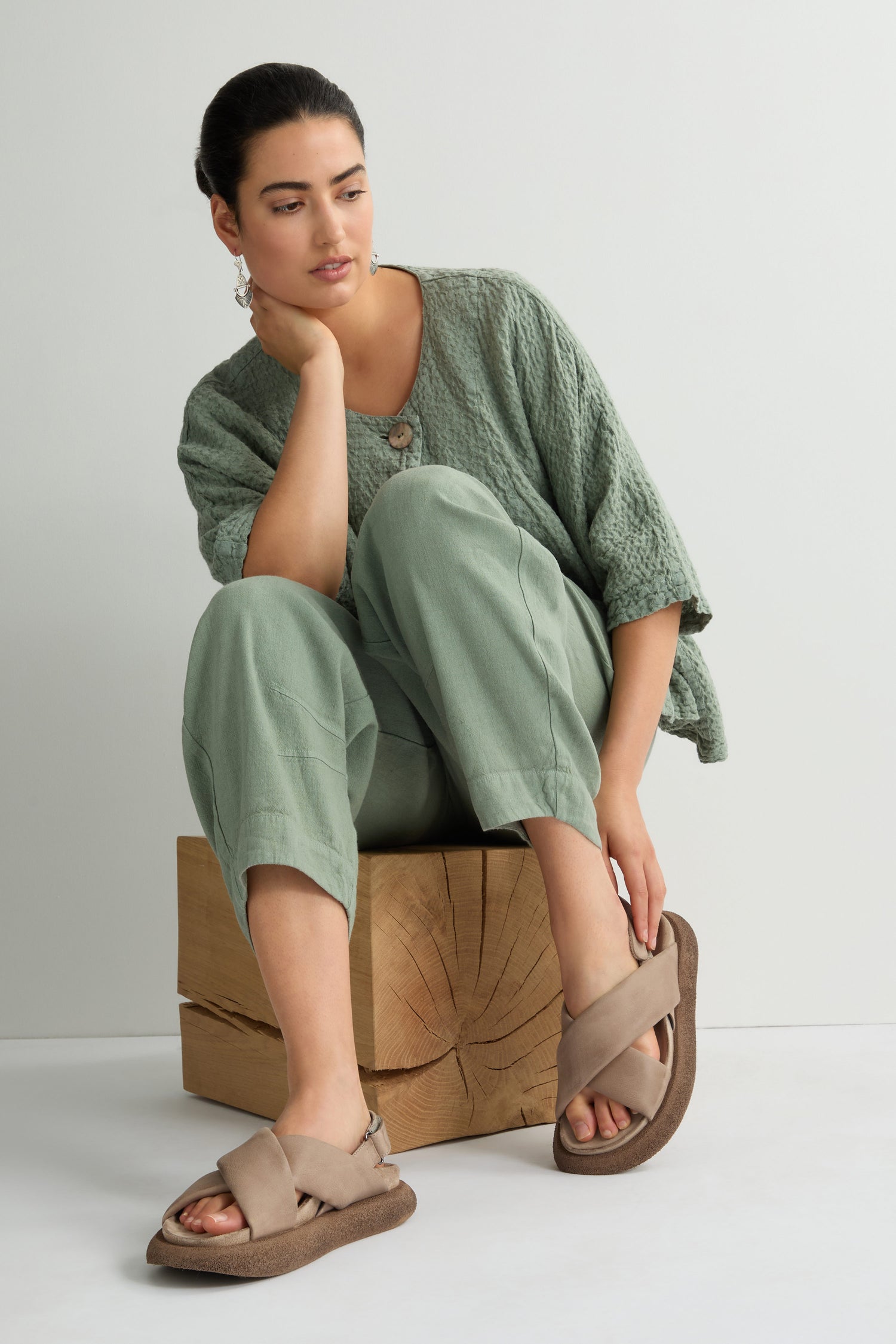 A woman in a textured green outfit and Crossover Leather Sandals featuring a cushioned footbed sits on a wooden block against a light background, showcasing Scandinavian craftsmanship.