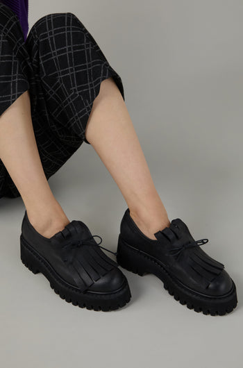 Person wearing Fringe Top Leather Brogues with thick, rugged soles and checkered black pants, sitting on a plain grey background.