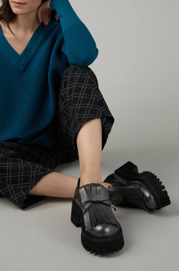 A person wearing a teal ribbed sweater, black and grey plaid pants, and Fringe Top Leather Brogues sits with one knee up and an arm resting on their knee against a plain background.