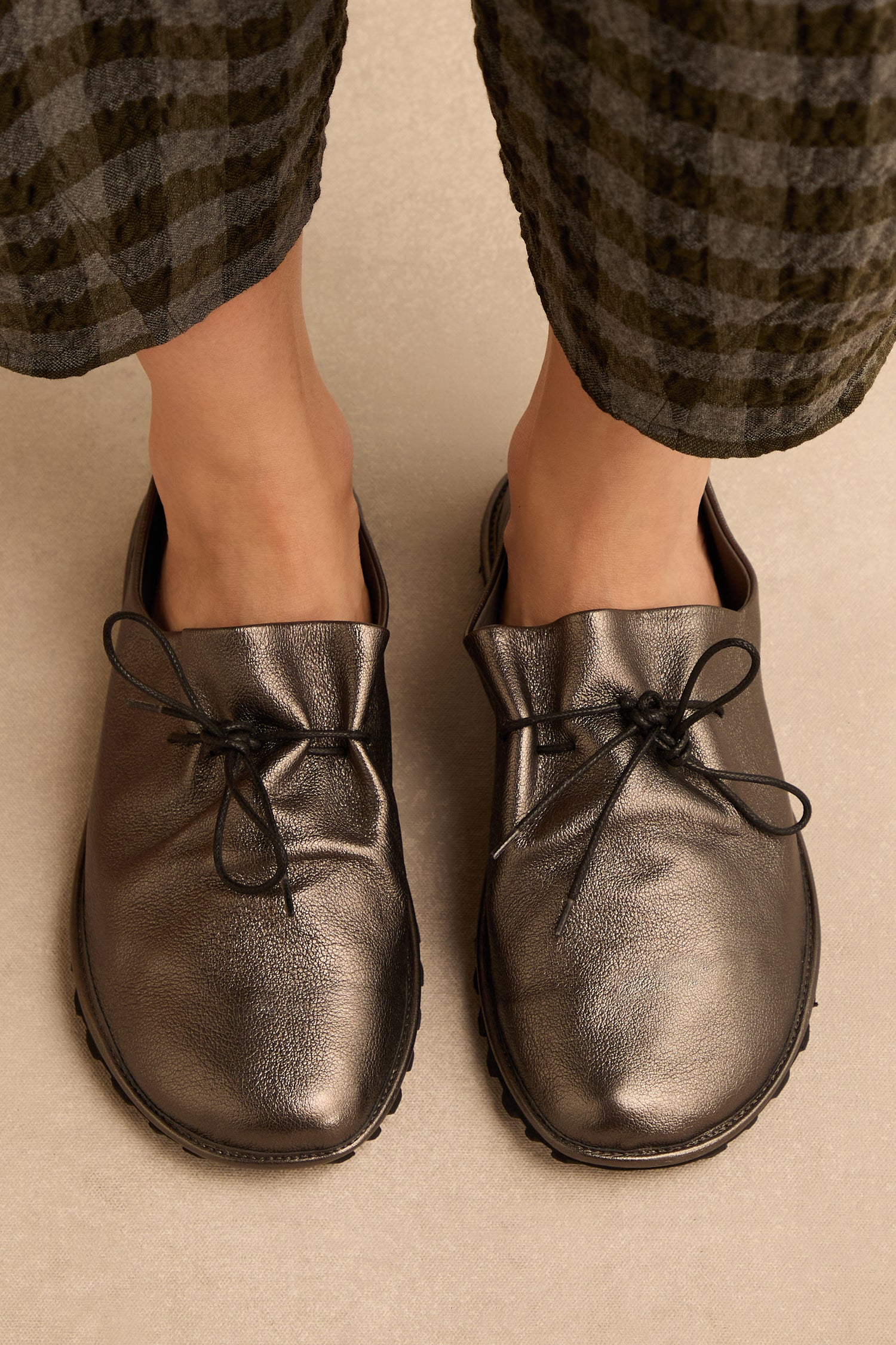 A person wears Hematite Crushed Leather Lace up Shoes, featuring a striking metallic leather finish, paired with patterned ankle-length pants and standing on a light-colored floor. Handmade in Italy, these statement shoes bring an artisanal touch to the modern ensemble.