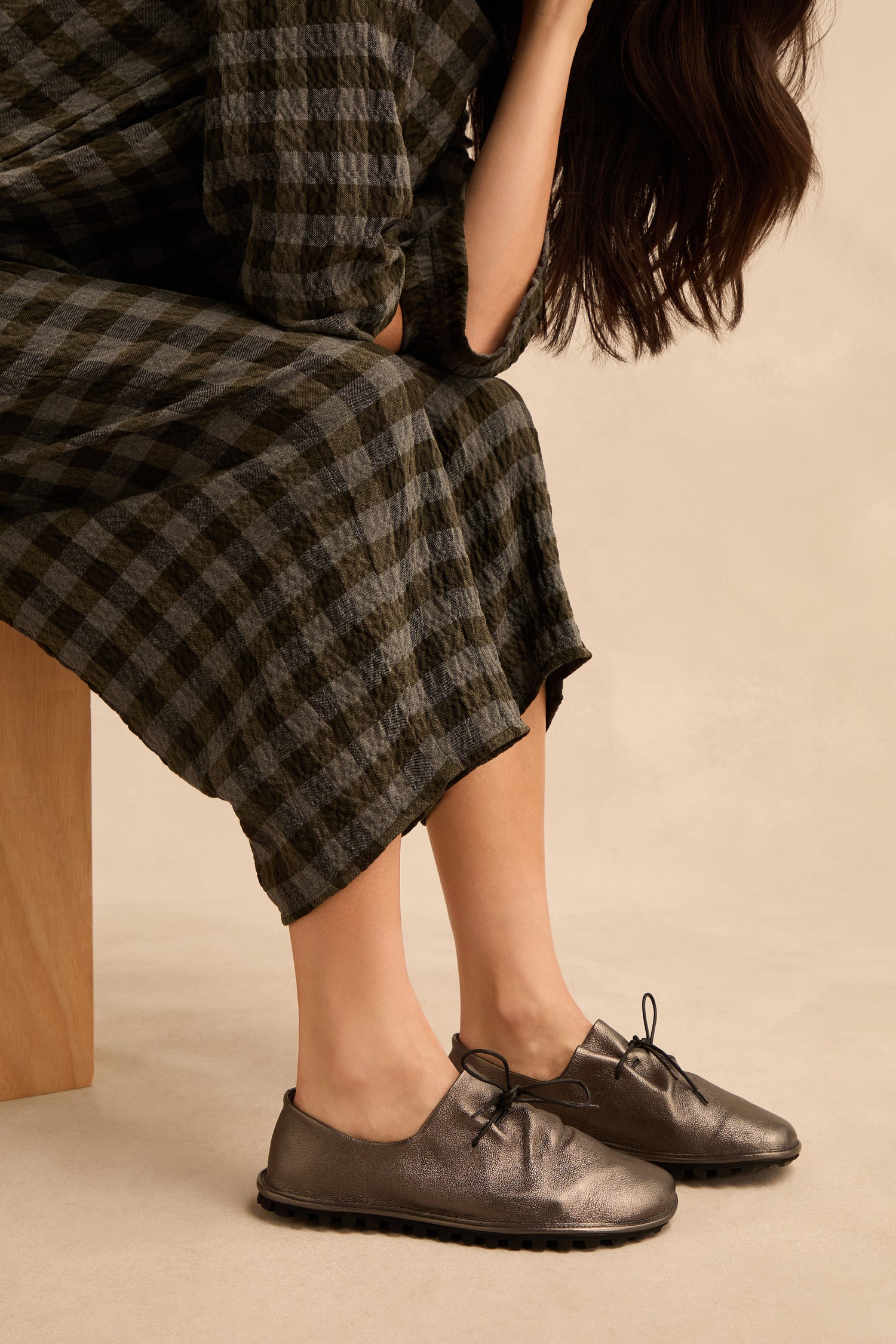 Sitting on a wooden bench, the person wears a long checkered dress and eye-catching Hematite Crushed Leather Lace up Shoes—featuring a metallic leather finish and handmade in Italy—with hair partially visible.
