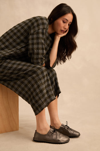 A woman in a checkered dress sits on a wooden block, resting her chin on her hand and gazing downward thoughtfully. She wears Hematite Crushed Leather Lace up Shoes that add a modern touch to her classic ensemble.