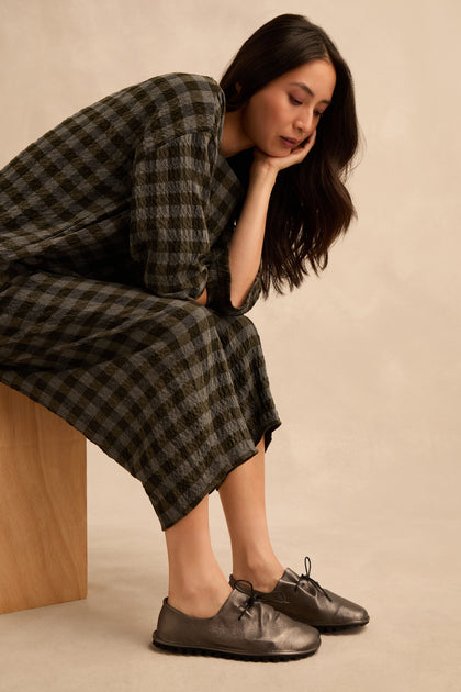 A woman in a checkered dress sits on a wooden block, resting her chin on her hand and gazing downward thoughtfully. She wears Hematite Crushed Leather Lace up Shoes that add a modern touch to her classic ensemble.
