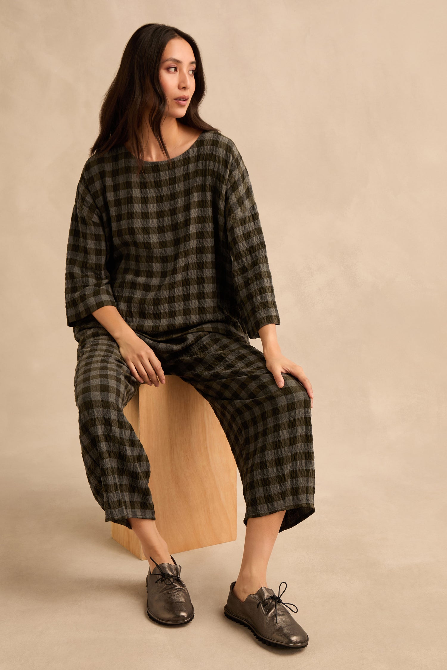 A woman sits on a wooden block wearing a matching dark green checked top and wide-leg trousers, her outfit completed with elegant Hematite Crushed Leather Lace up Shoes.