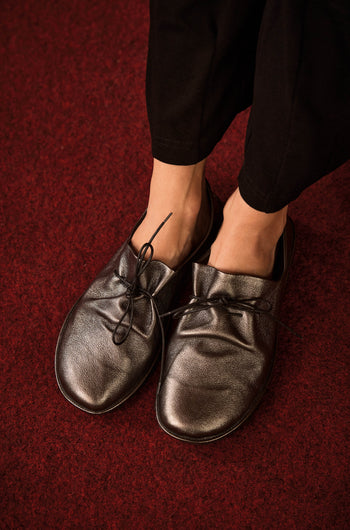 Wearing loose-fitting black Hematite Crushed Leather Lace up Shoes, handmade in Italy, a person stands on a dark red carpet.