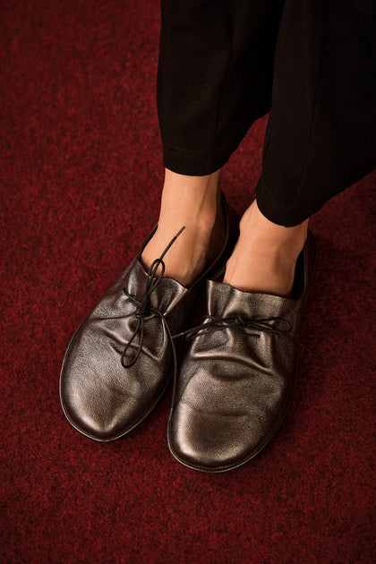 Wearing loose-fitting black Hematite Crushed Leather Lace up Shoes, handmade in Italy, a person stands on a dark red carpet.