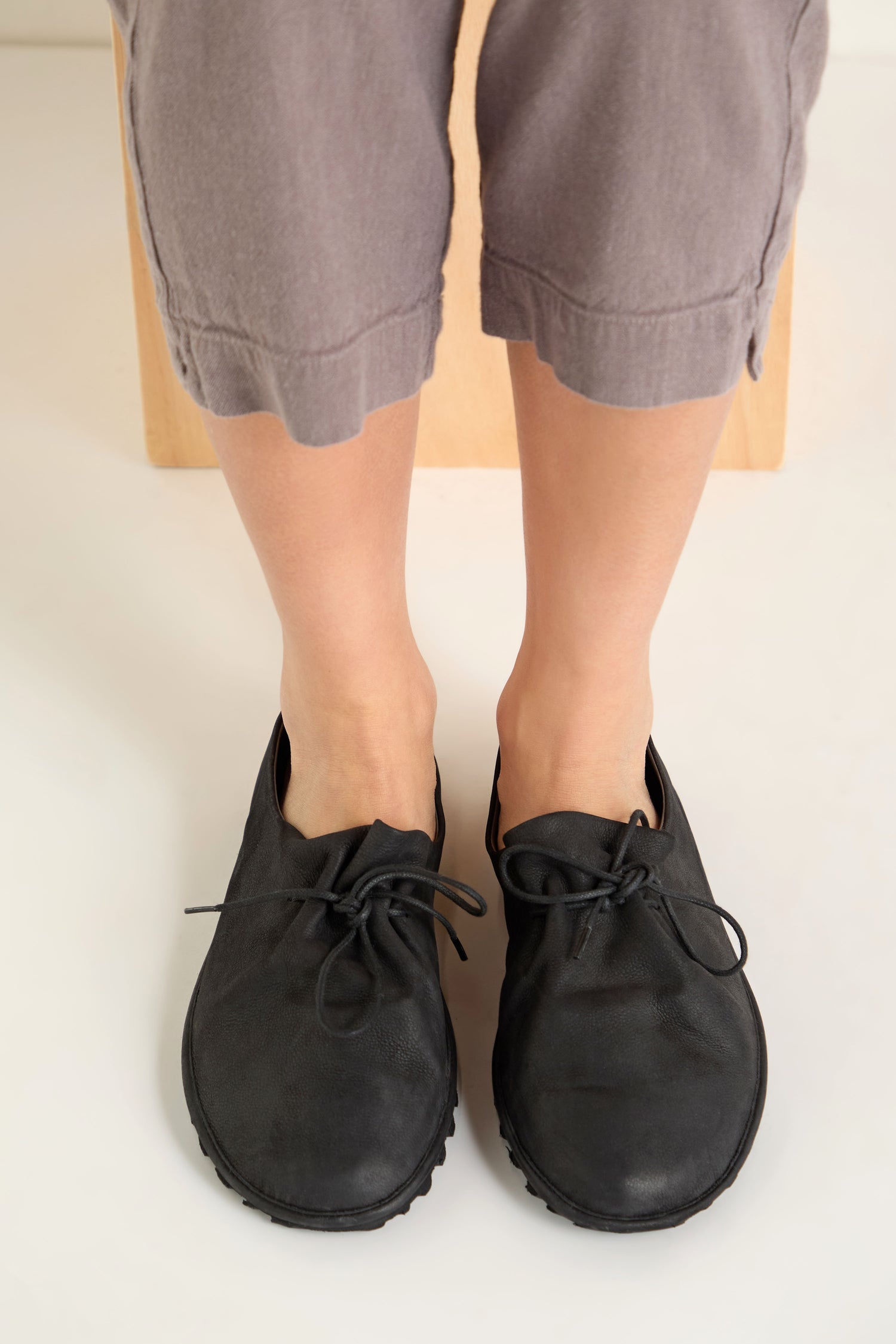 A person wears gray cropped pants and Crushed Leather Lace Up Shoes, handmade in Italy, while sitting with feet flat on a light-colored surface.