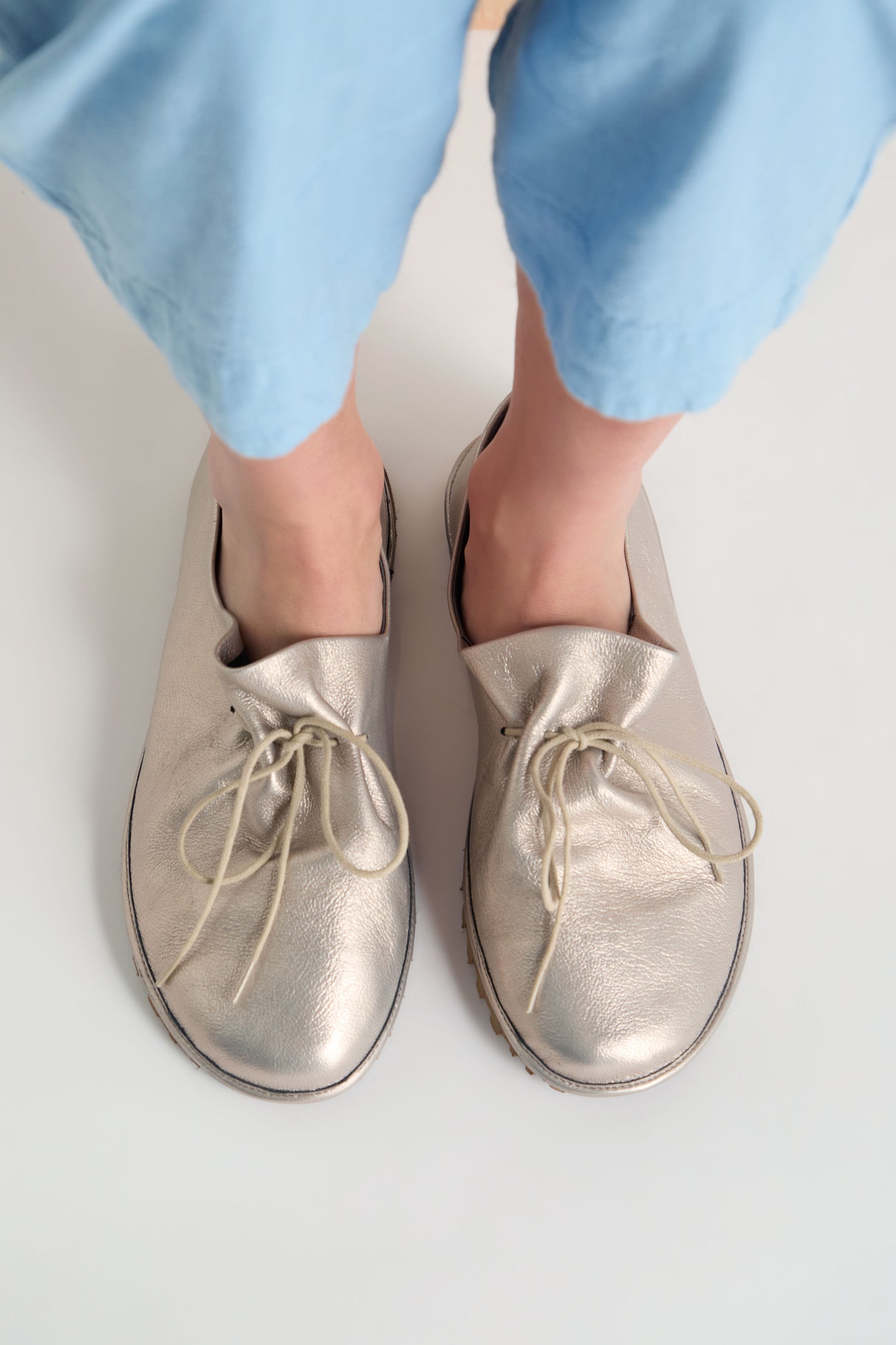 A person wearing light blue pants and oversized Crushed Leather Lace Up Shoes, handmade in Italy, stands on a white surface.