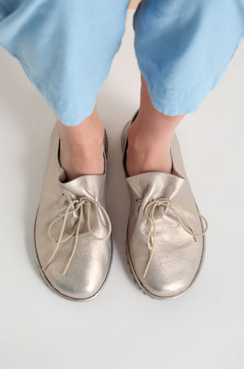 A person wearing light blue pants and oversized Crushed Leather Lace Up Shoes, handmade in Italy, stands on a white surface.