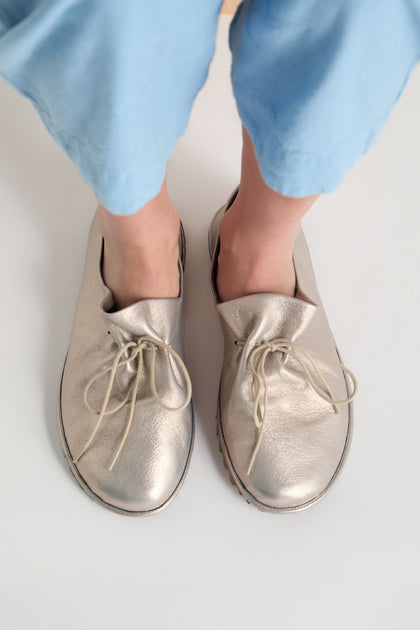 A person wearing light blue pants and oversized Crushed Leather Lace Up Shoes, handmade in Italy, stands on a white surface.