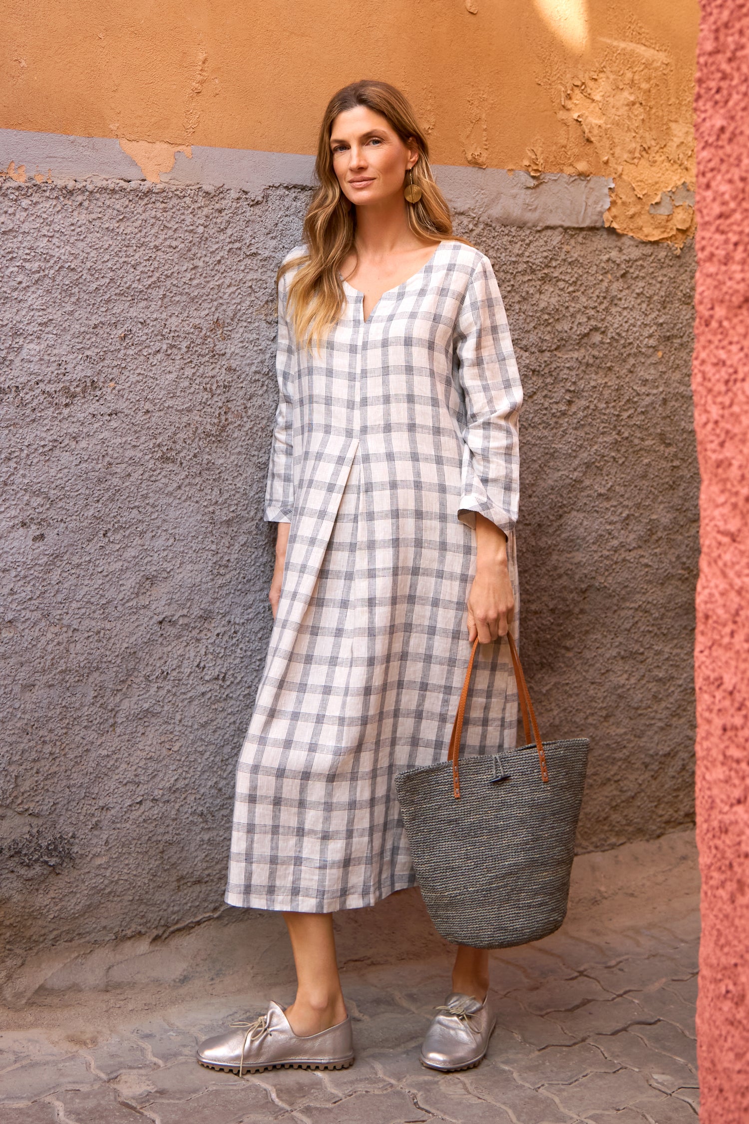 A woman stands against a textured wall wearing a long plaid dress and Crushed Leather Lace Up Shoes, holding a large woven tote bag.